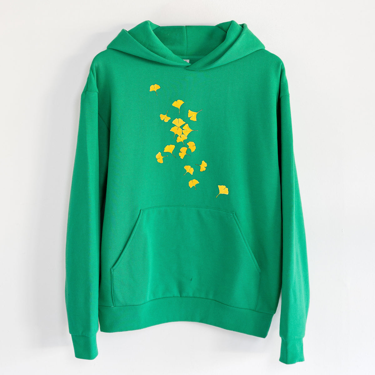 Falling Ginkgo Leaves - Statement Midweight Hoodie in Kelly Green, hanging on a white wall, with a front kangaroo pocket and no drawcords.