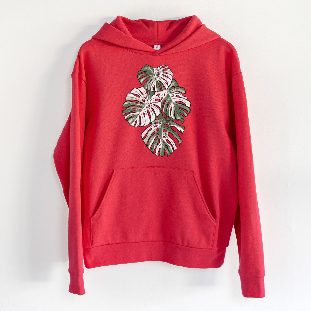 Variegated Monstera - Statement Midweight Hoodie in Cherry Red, hanging on a white wall, with a front kangaroo pocket and no drawcords.