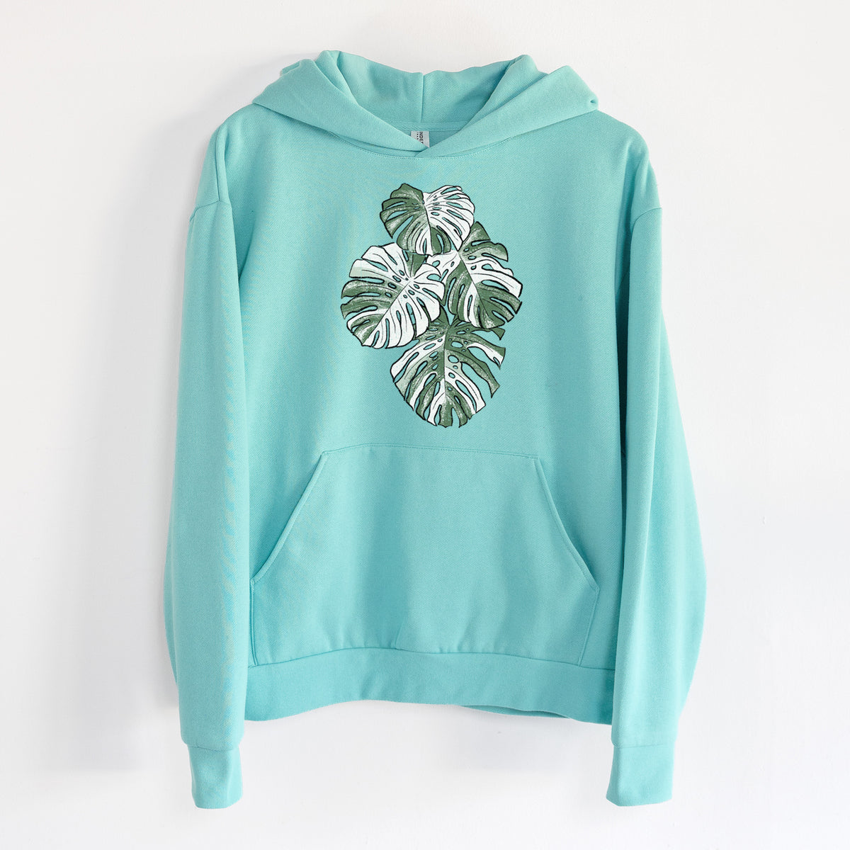 Variegated Monstera - Statement Midweight Hoodie in Seafoam, hanging on a white wall, with a front kangaroo pocket and no drawcords.