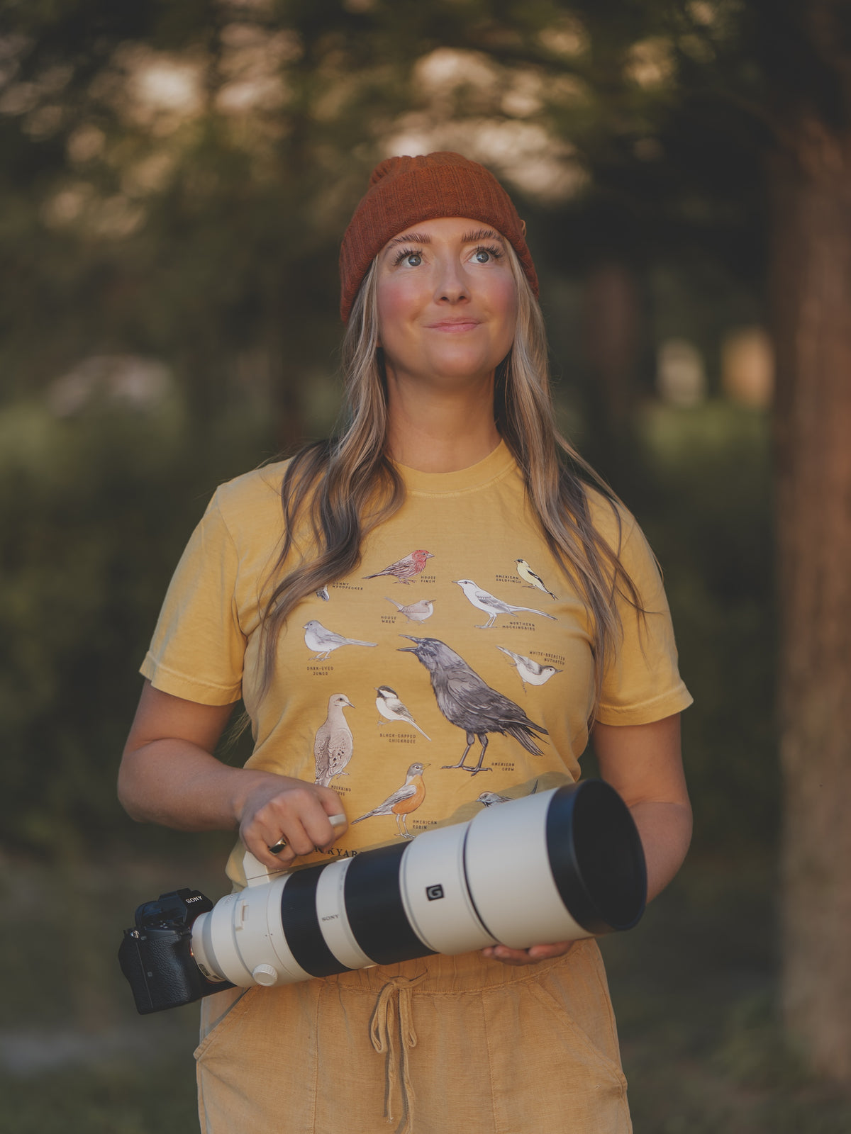 A woman wearing a mustard yellow graphic t-shirt with bird illustrations, a rust-colored beanie, and tan pants, holding a large camera lens outdoors in a wooded setting.